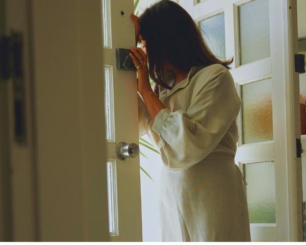 Person in a light-colored outfit standing in a doorway with a plant in the background