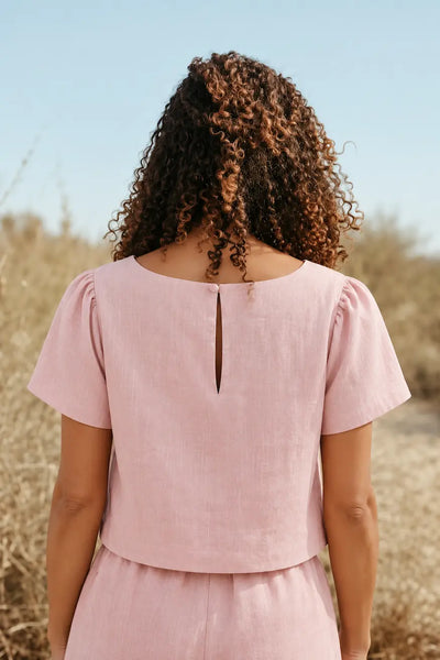 A woman viewed from behind wearing a light pink linen short-sleeve top with puff sleeves and a small keyhole cutout at the back